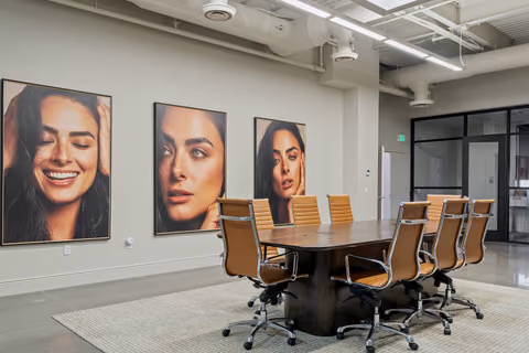Conference room with a dark wood oval table, tan leather office chairs, and three large framed portrait photographs on a grey wall, with exposed white ductwork overhead and a glazed corridor at right.