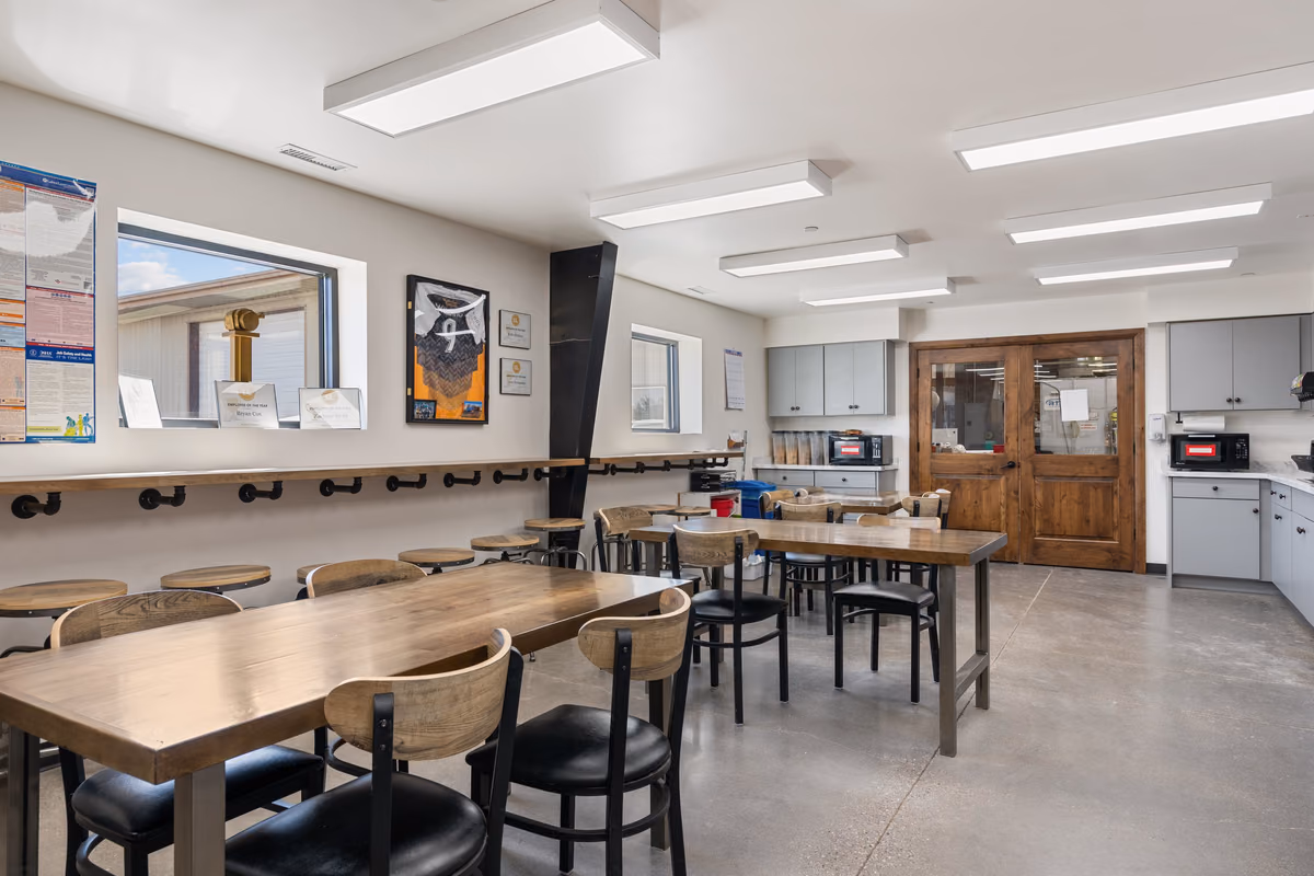 Employee break room with wood-topped tables, black chairs with wood backs, a pipe-bracketed counter with stools under a window, framed recognition plaques on the wall, and wooden double doors.