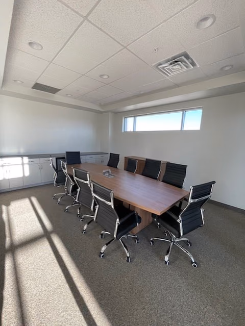 Conference room with a long wood-topped table, black leather office chairs, a high ribbon window, and white cabinets along one wall.