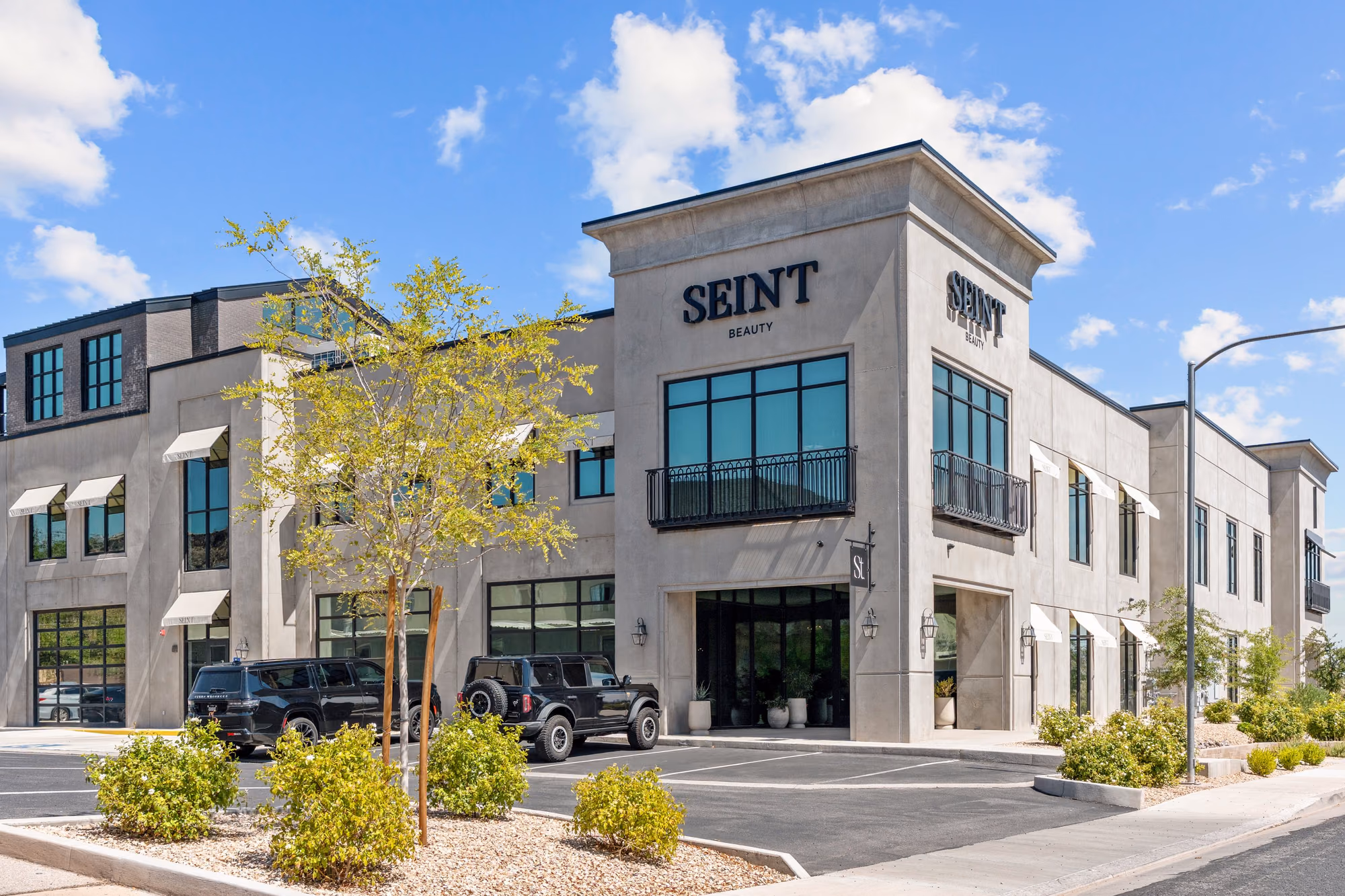 Corner of a two-story grey concrete office building with black steel-framed windows, wrought-iron balconies, cream awnings, and black SEINT BEAUTY lettering above the entrance, under a blue sky with clouds.