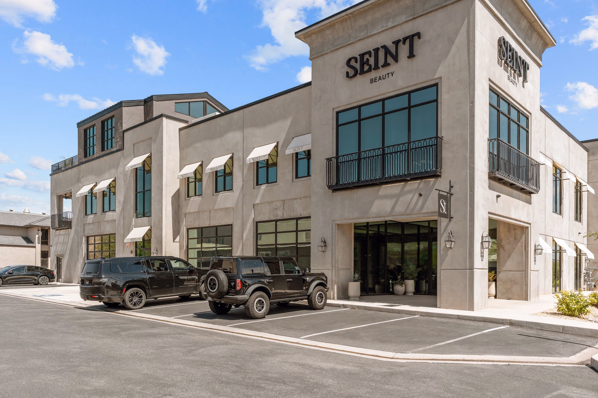 Street-level view of the grey concrete office building with the recessed glass entrance, black SEINT BEAUTY lettering, iron balconies, cream awnings, and two black SUVs parked in front.