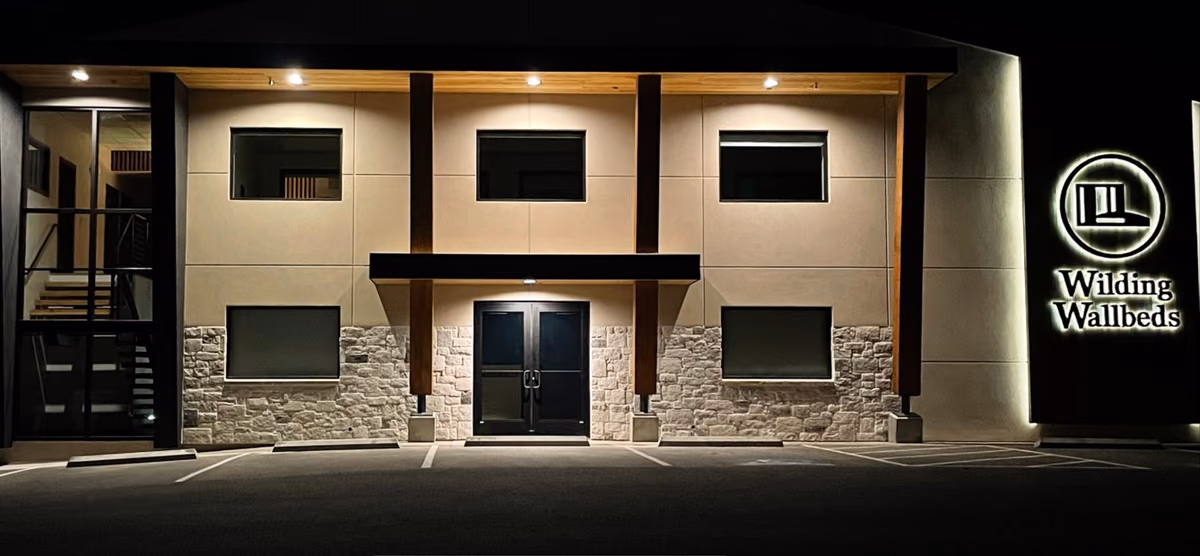 Front of the building at night, warmly lit from above, with cedar posts, a stone base, and the illuminated Wilding Wallbeds logo and name on the sign wall at right.