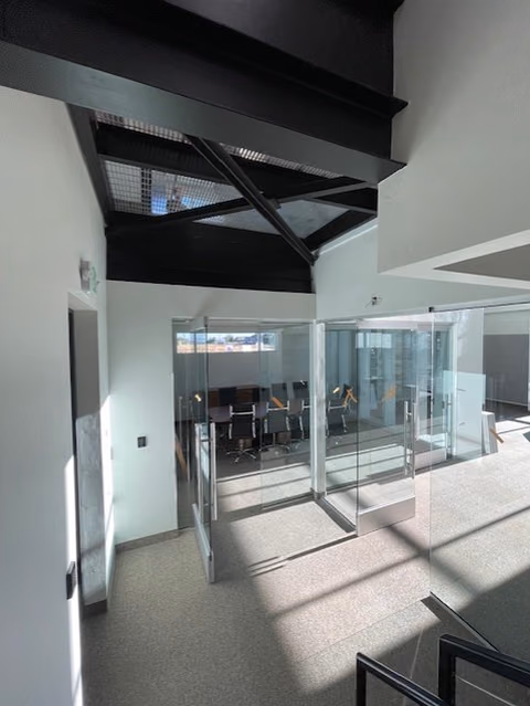 Glass-walled conference room seen through glass doors beneath a black steel skylight frame, with bars of sunlight across a speckled carpet floor.