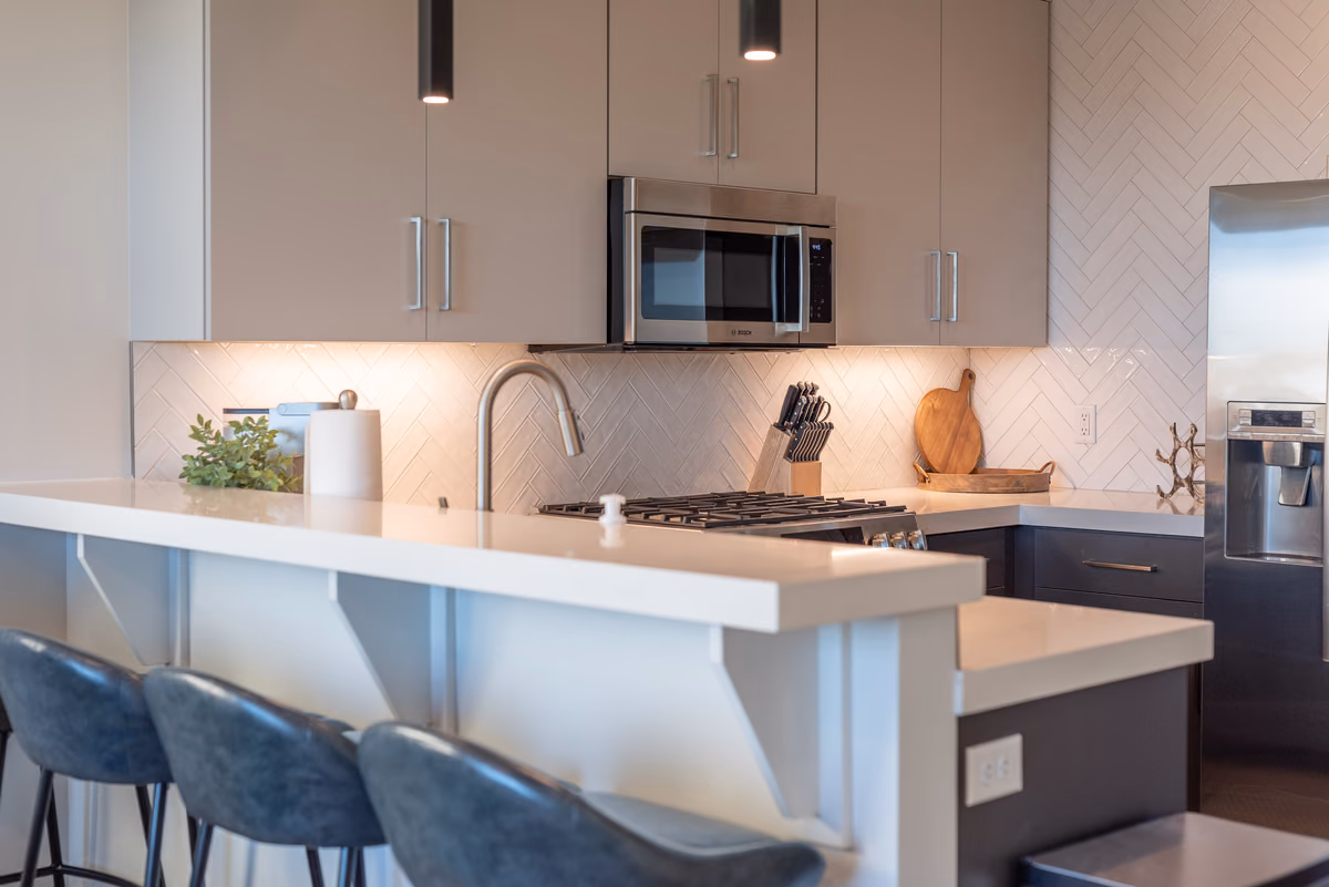 Kitchen counter with a white stone bar top, three blue leather stools, a herringbone tile backsplash with under-cabinet lighting, a gas cooktop, and a stainless microwave.
