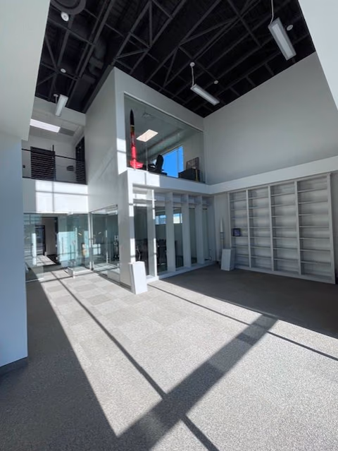 Double-height room with a black steel roof structure, tall windows, white built-in shelving along the walls, and a glass-fronted room on an upper level at the back.