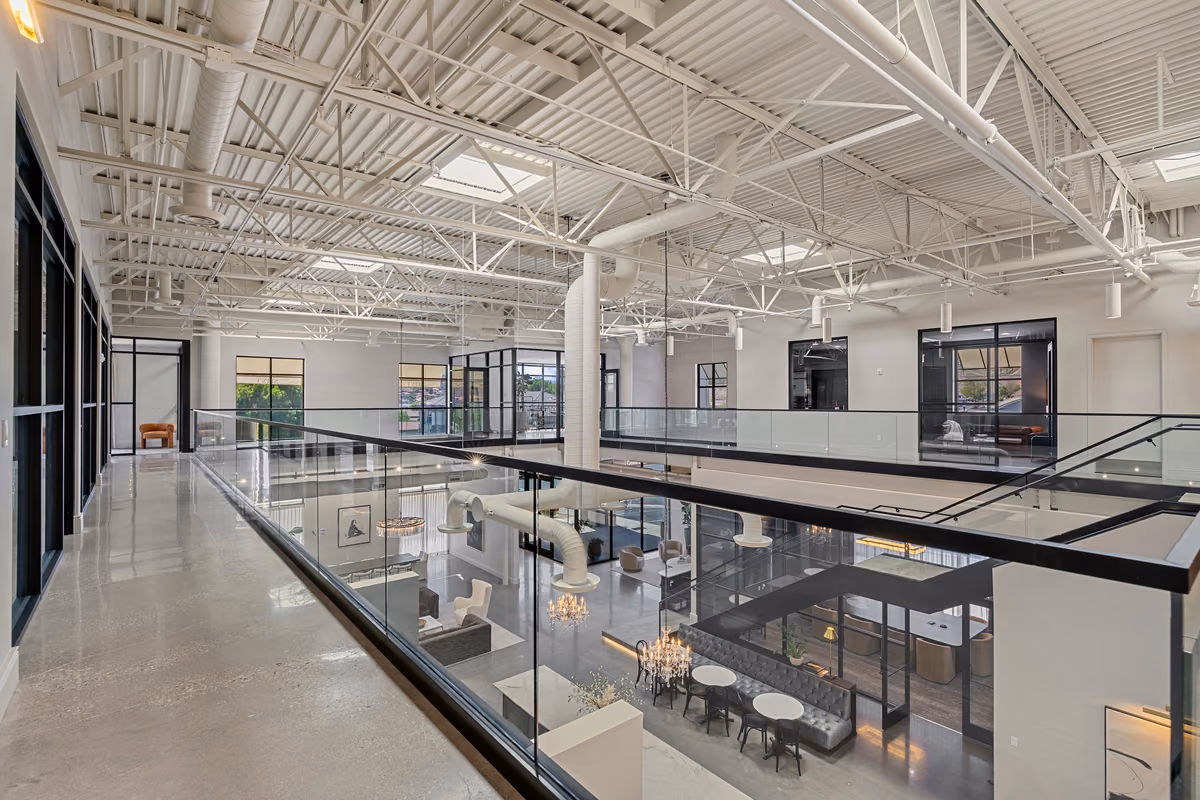 Mezzanine corridor with a polished concrete floor beside a glass guardrail, looking down on the café and banquette, under white steel trusses, skylights, and round ductwork.