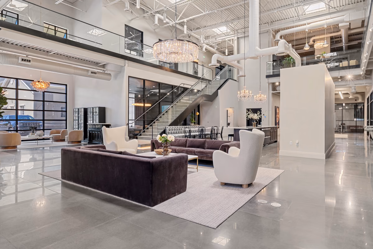 Ground-floor lounge with a purple-grey velvet sofa, white boucle wing chairs, a crystal chandelier, a glass-railed stair, and the café behind, on polished concrete.